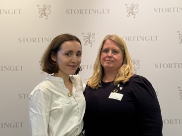 To kvinner står foran en hvit vegg som det står stortinget på, og som har stortingets logo på seg. Kvinnen til venstre er leder av Norsk medisinstudentforening, Elise Skimmeland. Hun har brunt hår til haka som er bak ørene, og hun smiler forsiktig. Hun har hvit skjorte på seg. Kvinnen til høyre er  leder i Yngre legers forening, Ingeborg Henriksen. Hun har blondt hår til nedforbi skuldrene, har på seg briller og sort genser. Hun ser alvorlig ut.  
