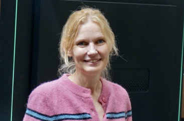 Kvinne, lyst hår, rosa strikkejakke med blå og svart stripe. Mørk bakgrunn.