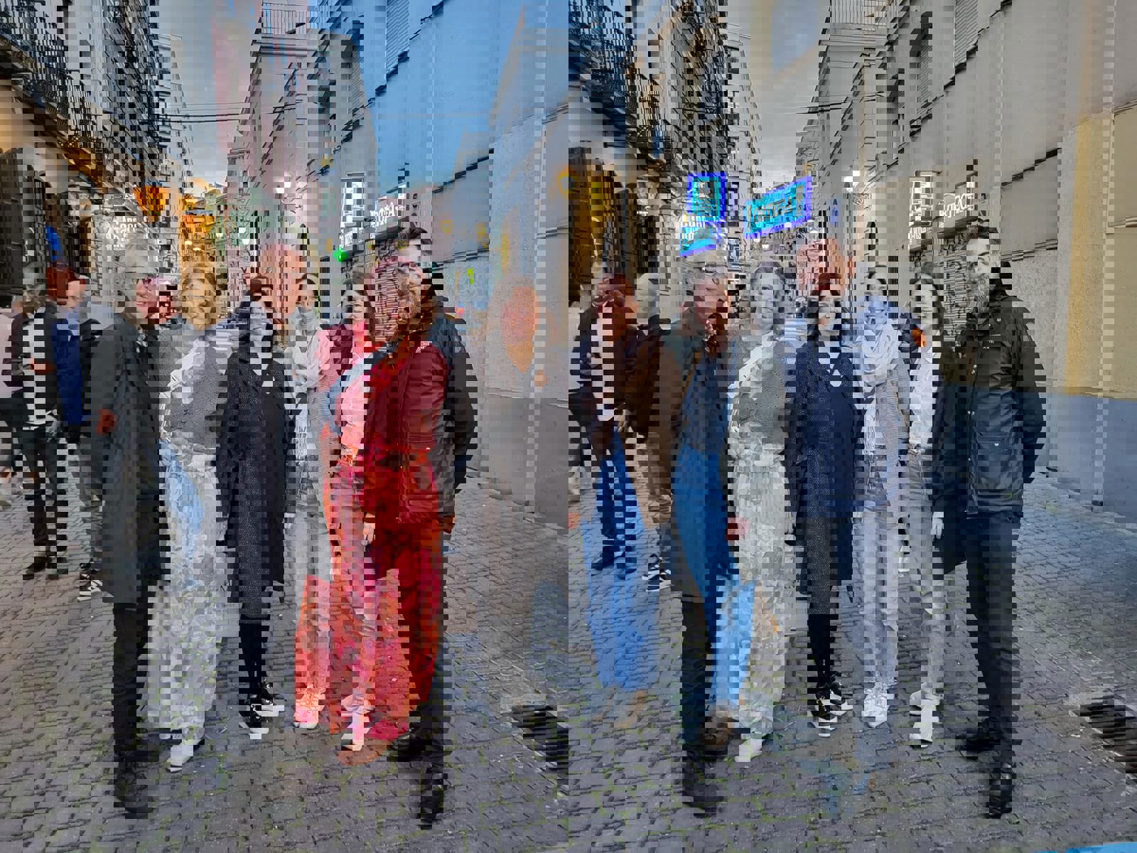 Seks mennesker i en gate. Mann i mørke klær, kvinne i rødlig klær, kvinne i mørke klær, to kvinner i lyse topper og olabukser, mann i mørke klær-