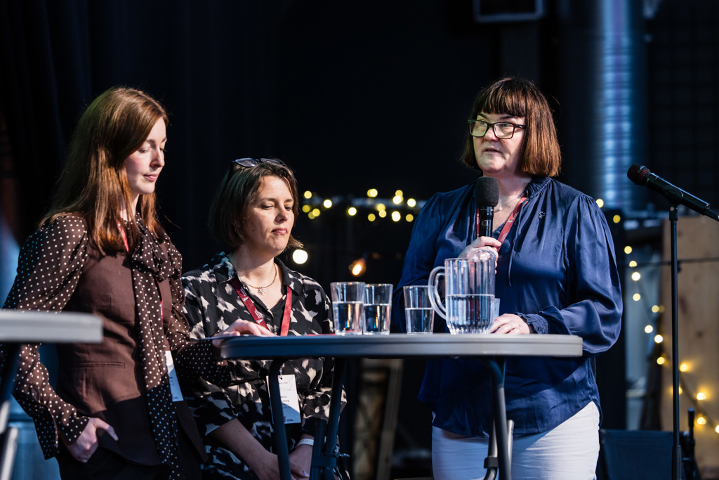 Carolin Sparchholz, nestleder i Yngre legers forening, Marie Skontorp, nestleder i Overlegeforeningen og nestleder i Allmennlegeforeningen, Else Worren Nygård, deltar i en panelsamtale på en scene rundt et høyt bord. Nygård står og holder en håndholdt mikrofon, mens de to andre står og sitter ved siden av. På bordet foran dem står flere gjennomsiktige glass med vann og en vannkanne. Bakgrunnen er mørk, med små punktlys og synlig scenebelysning som gir en tydelig arrangementsstemning.