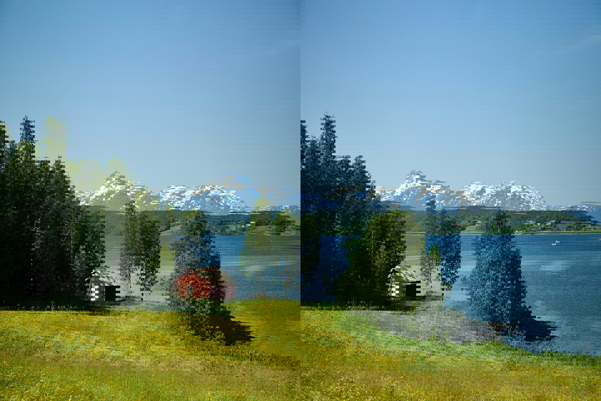 Blå himmel, snødekte fjell, grønt gress, hav og en liten rød hytte. Illustrasjonsbilde.