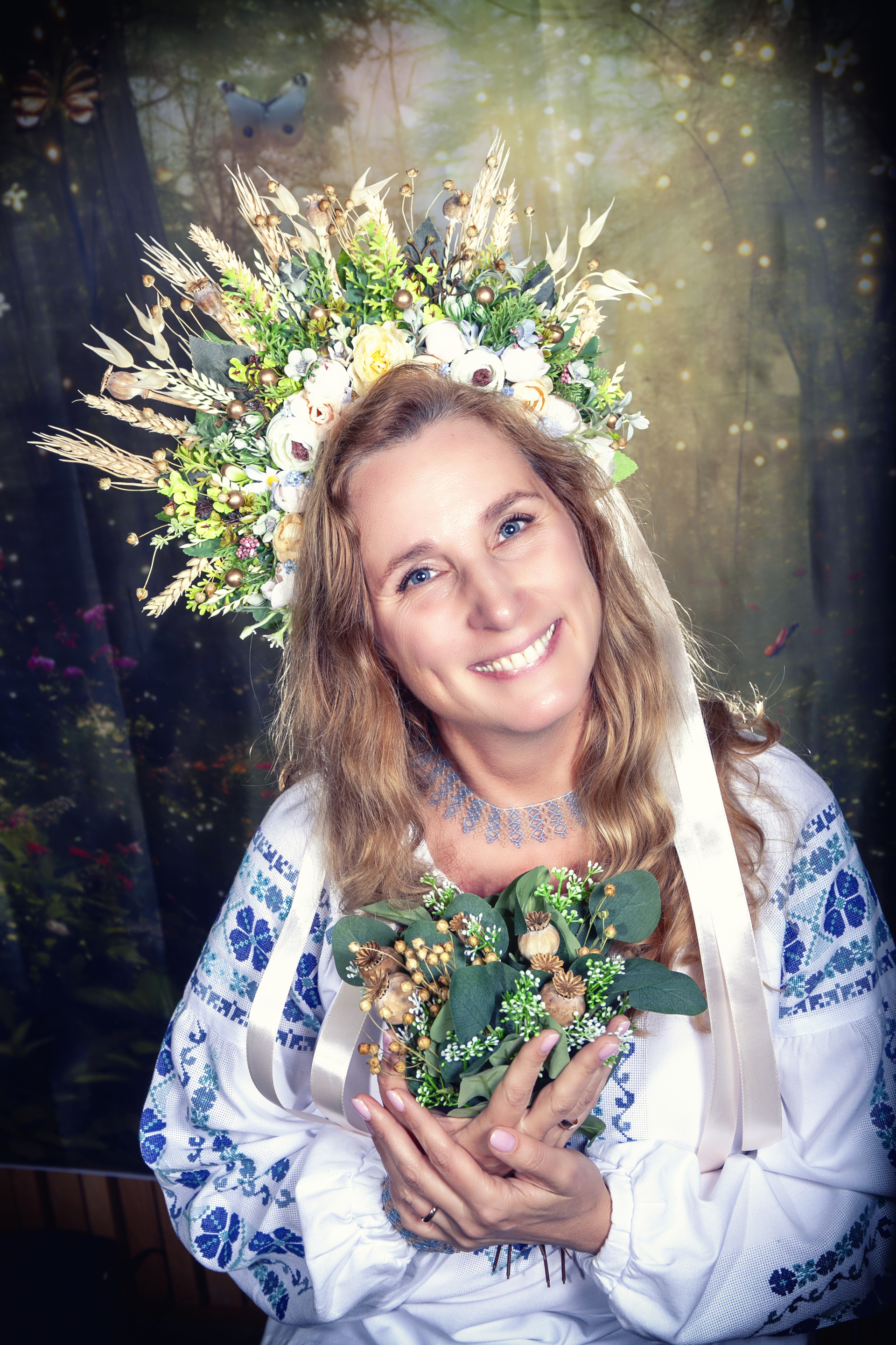 Smilende kvinne med blomsterpynt i håret og i armene. Lyst hår. Hvit bluse med blå broderier.