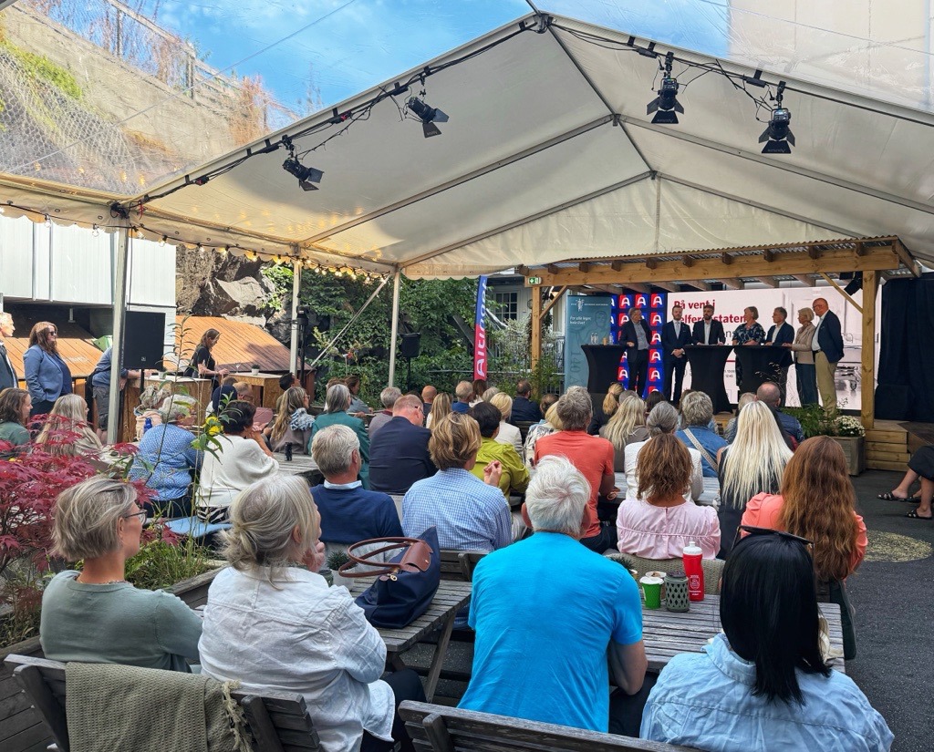 Publikum sitter ved bord og benker under et telt og følger en paneldebatt på en scene i bakgrunnen.
