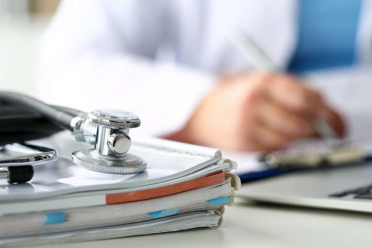 Lege som sitter ved en pult og skriver, med bøker, pc og et stetoskop i forgrunnen. Foto: Istockphoto.com