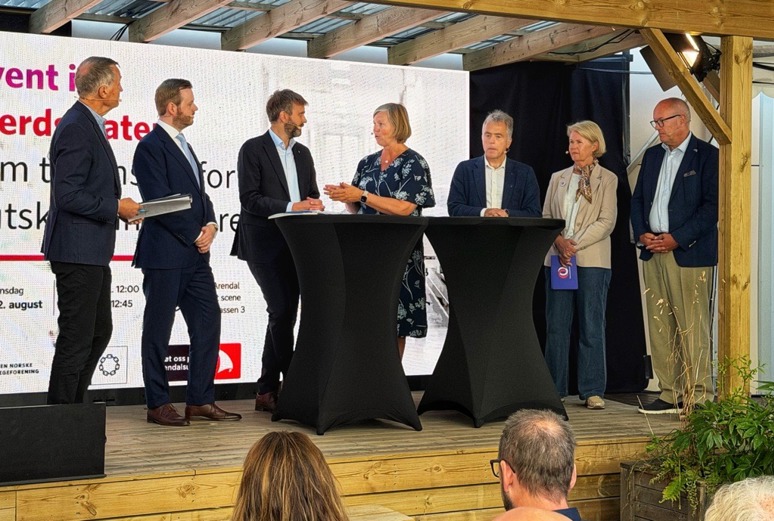 Seks personer deltar i en paneldebatt på en scene. To ståbord står foran panelet, og en storskjerm med arrangementstittelen vises i bakgrunnen.