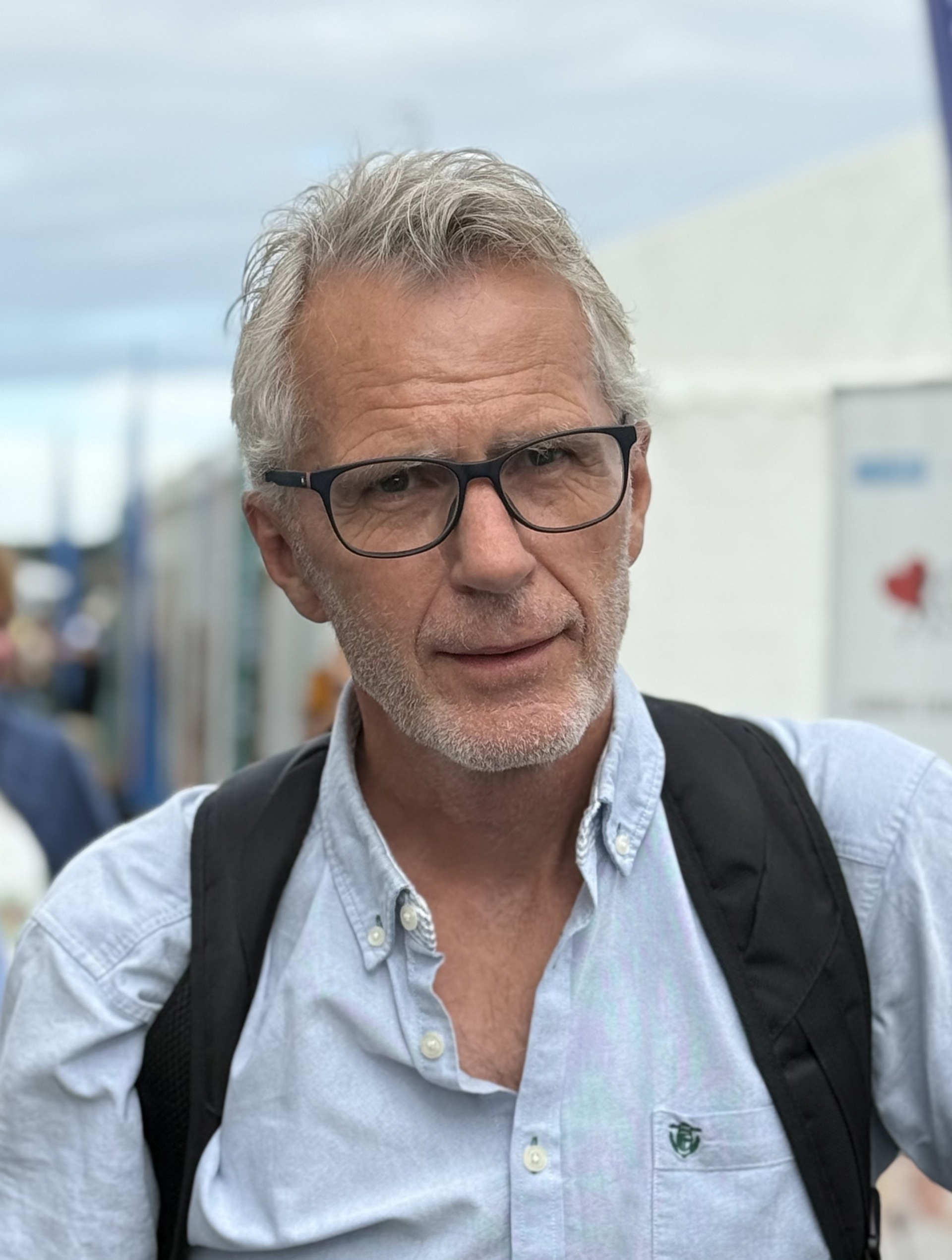 Mann, grått hår og skjegg, sekk på ryggen, lyseblå skjorte. Briller. Himmel i bakgrunnen.