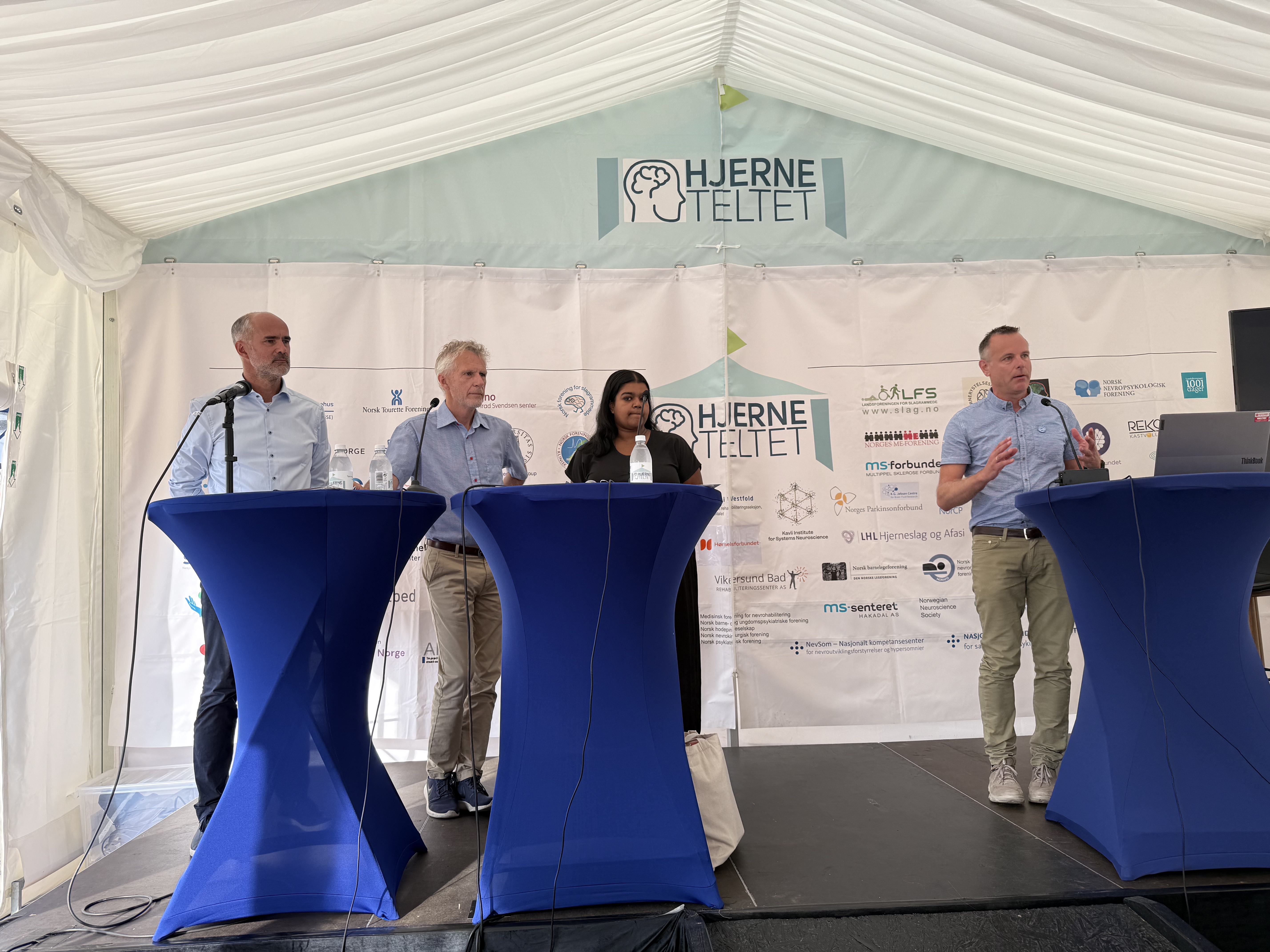 Tre menn i lysebl&aring; skjorter og en kvinne i m&oslash;rk jakke bak bl&aring; talerstoler p&aring; en scene.