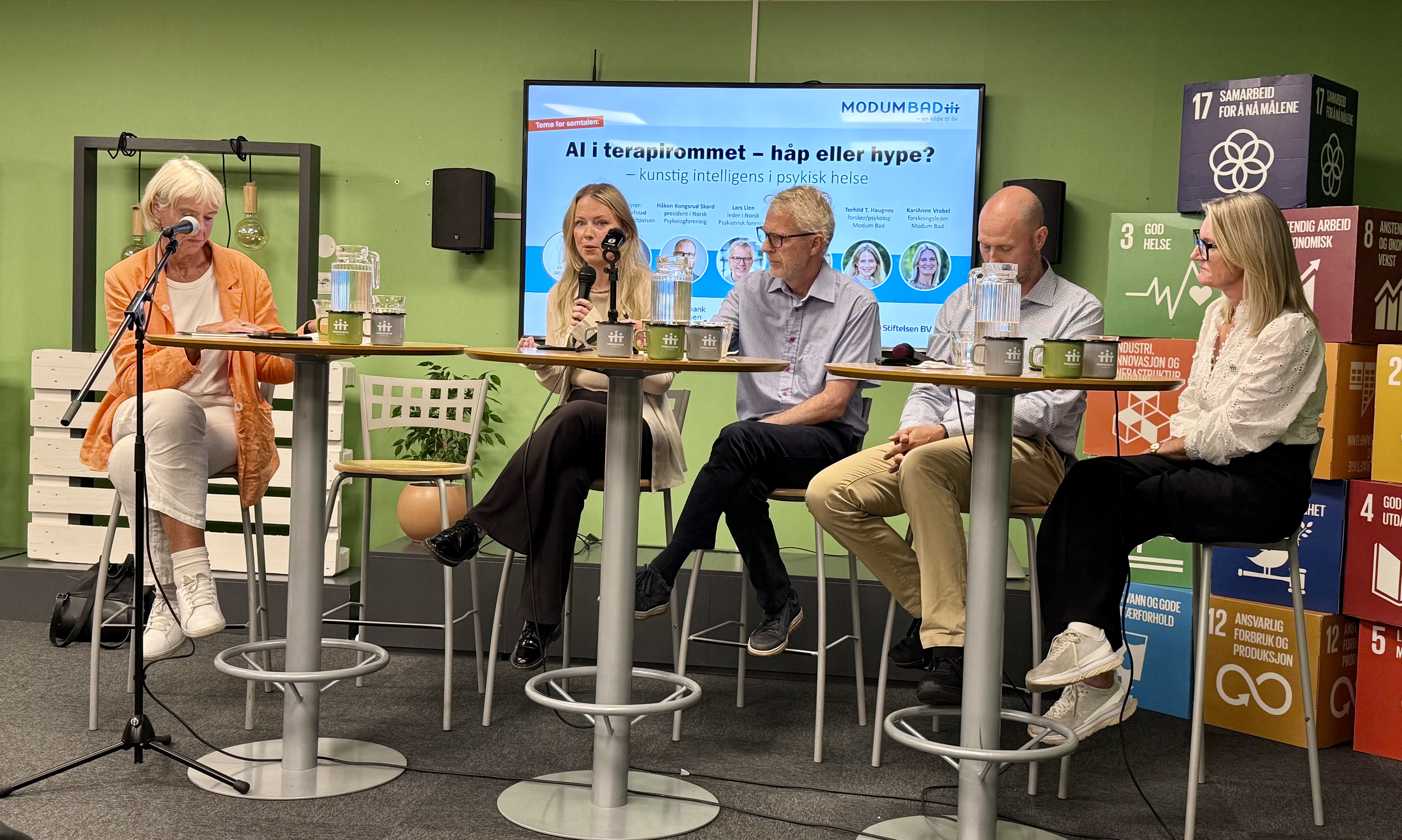 Tre kvinner og to menn p&aring; barkrakker bak sm&aring; runde bord p&aring; en scene