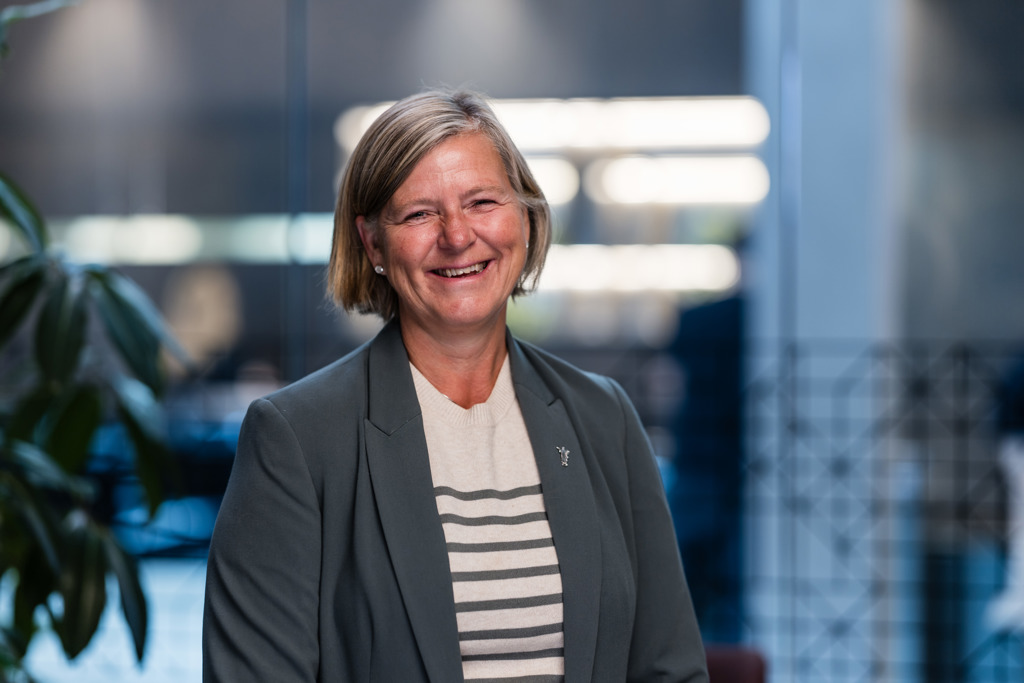 Presidenten i Legeforeningen, Anne-Karin Rime. Hun har kort, lyst hår til haka. Håret er gredd i sideskill og ligger bak ørene. I øreflippene har hun perler. Hun smiler, og har på seg en grå blazer med en hvit genser med grå striper inni. Bakgrunnen er utydelig, men viser et kontorlandskap med planter i venstre hjørne.