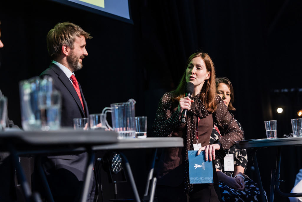Carolin Sparchholz, nestleder i Yngre legers forening står ved et panelbord på en scene og holder en mikrofon mens hun snakker. Jan-Christian Vestre står ved siden av og vender seg mot taleren, og flere personer er delvis synlige i bakgrunnen. På bordet står flere glass med vann og en gjennomsiktig vannkanne. Bakgrunnen er mørk med scenelys og en projeksjonsskjerm øverst i bildet.