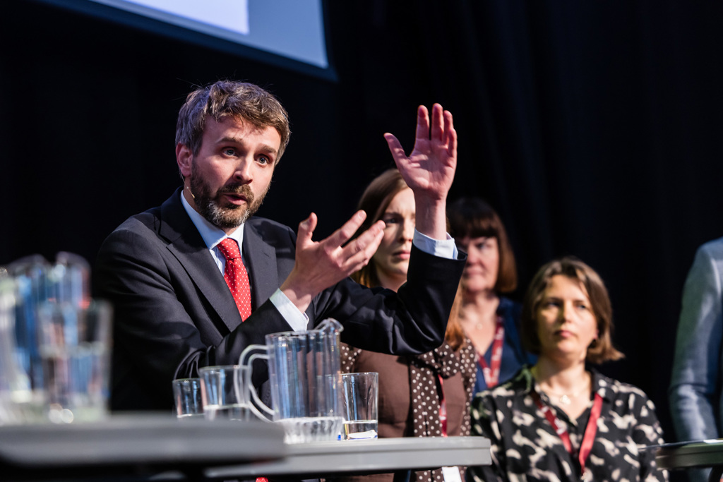 Helseminister Jan-Christian Vestre i dress og slips står ved et panelbord på en scene og snakker mens hendene løftes i en forklarende bevegelse. Tre kvinner sitter ved siden av og bak på samme rad, de er uklare. Foran dem står gjennomsiktige vannkanner og drikkeglass. Scenen er svakt opplyst med mørk bakgrunn og en projeksjonsskjerm øverst i bildet.