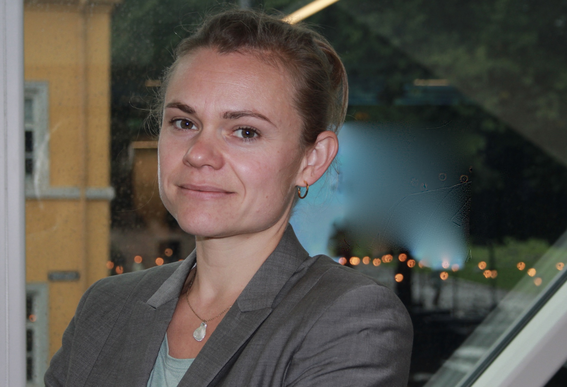 Kvinne med blondt hår. grå jakke og lys blå topp. I bakgrunnen et vindu.