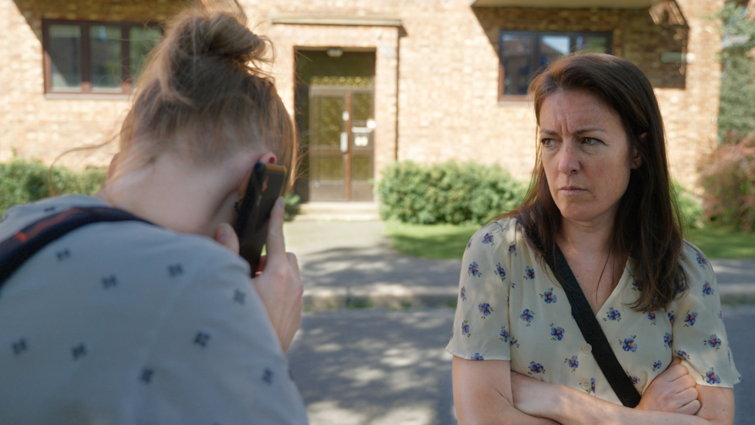 To kvinner ute på gata. rød bygning bak. grønt gress.