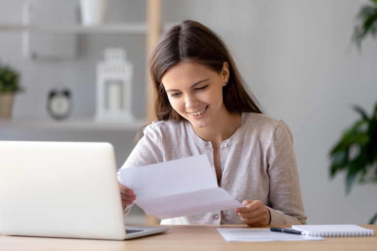 Kvinne som smiler mens hun leser et ark foran en pc. Foto: Istockphoto.com
