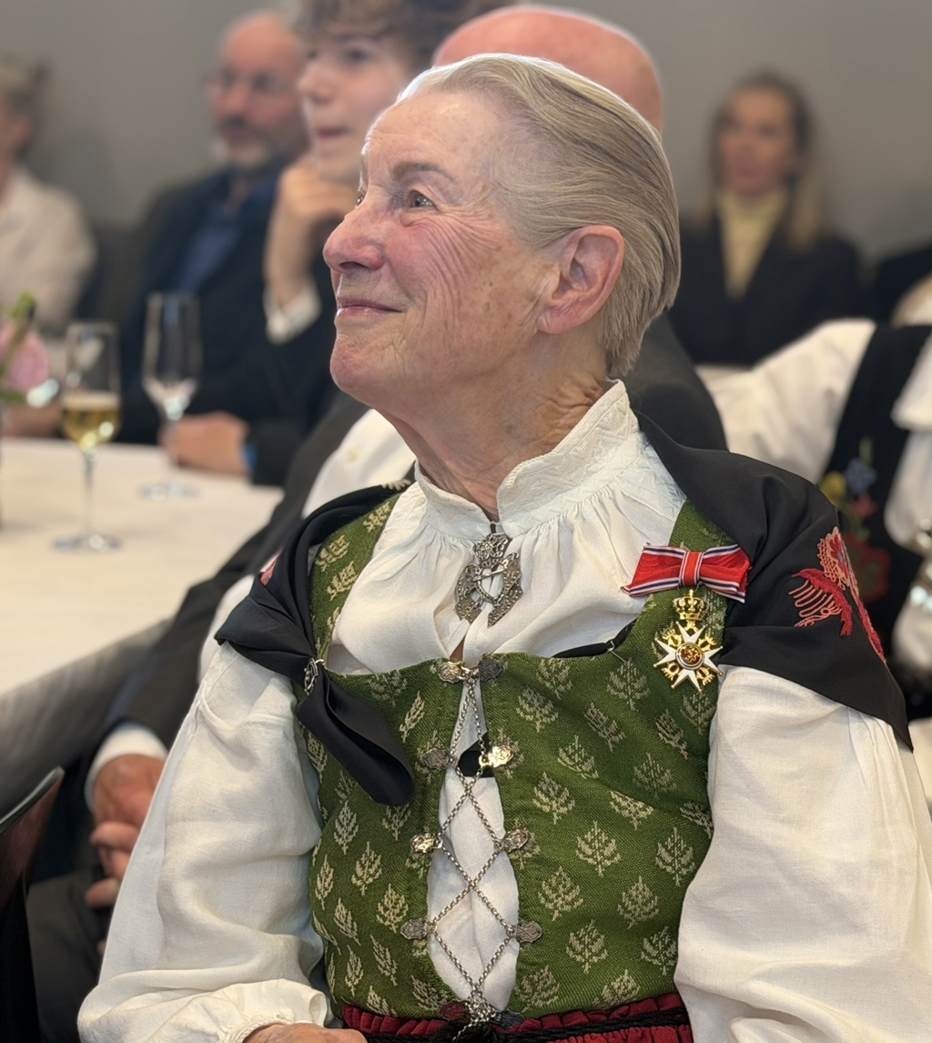 Kirkengen er ikledd bunad og sitter ved et dekket bord og bærer en orden festet på brystet. I bakgrunnen sitter andre gjester, og det står blomster og glass på bordet.