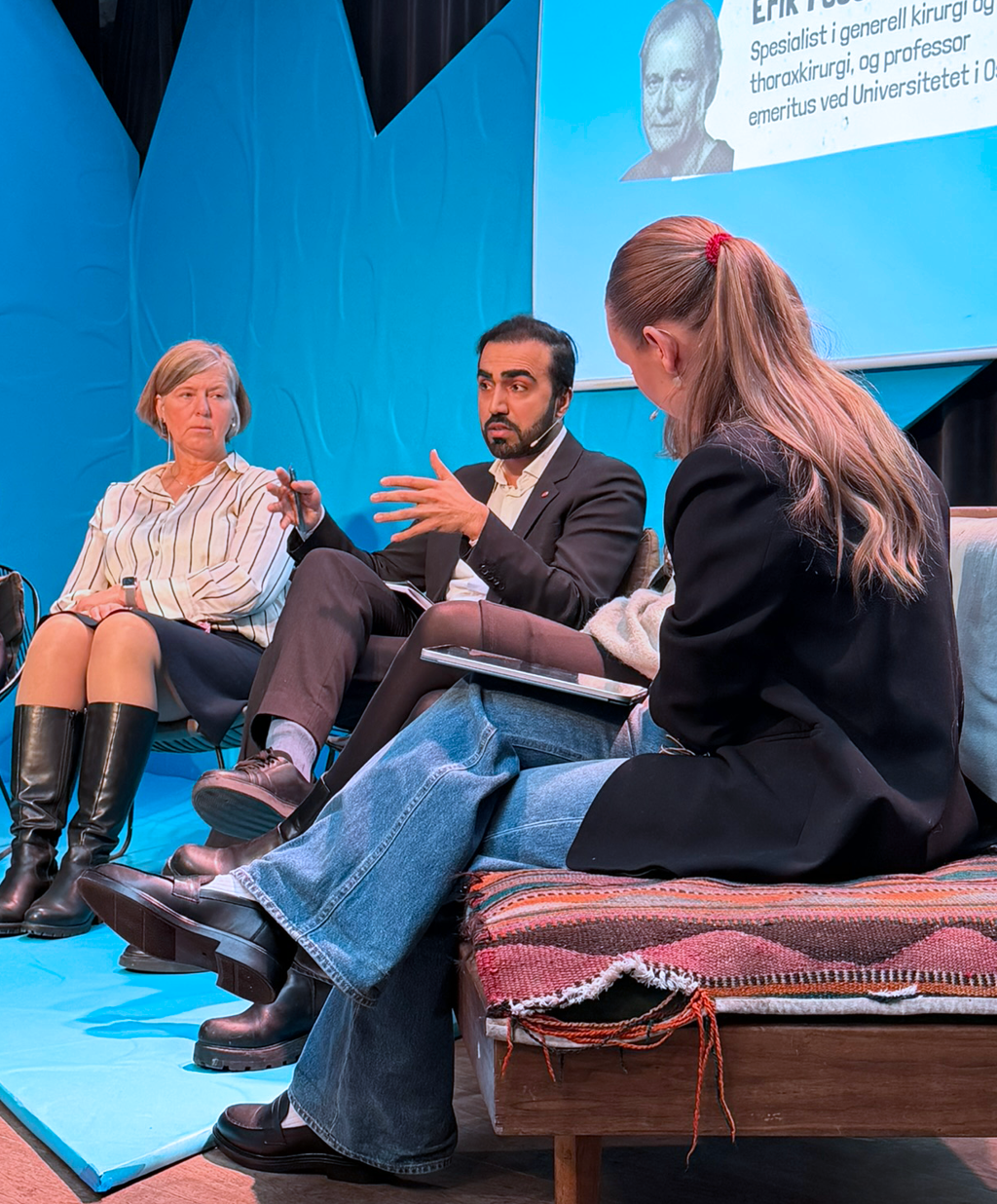 Du ser (f.v.) Anne-Karin Rime, Usman Mushtaq og Jeanett Myren. De sitter foran en blå bakgrunn. Rime har lyst, kortklippet hår til rett nedforbi ørene. Hun har på seg hvit skjorte med sorte striper, grått skjørt til knærne og sorte støvletter til rett under kneet. Hun har hendene i fanget og ser på Mushtaq. Han prater og holder hendene ut foran seg, med sprikende fingre. Han har sort hår og skjegg, mørk dress og hvit skjorte. Han har lyseblå sokker som skimtes, og sorte dressko. Han har beina i kors. Myren ser vi ryggen på og ansiktet delvis i profil. Hun har langt, lysebrunt hår i hestehale (rød strikk), sort dressjakke og blå, løse jeans. Hun har på seg sorte sko. Hun har beina i kors.  