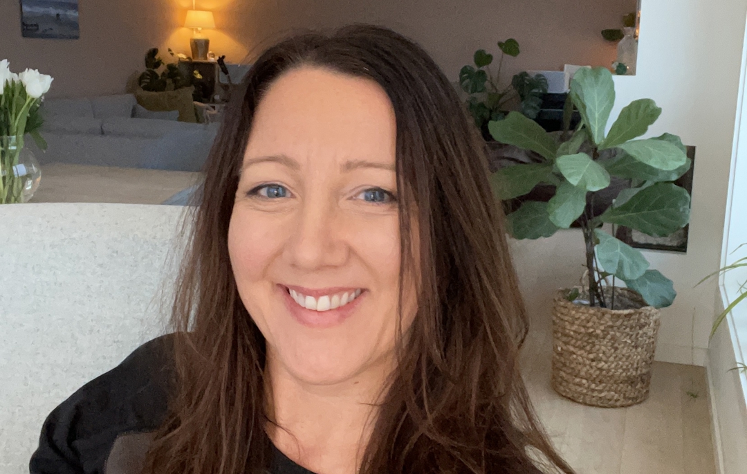 Kvinne, smilende, langt brunt hår. Stue i bakgrunnen