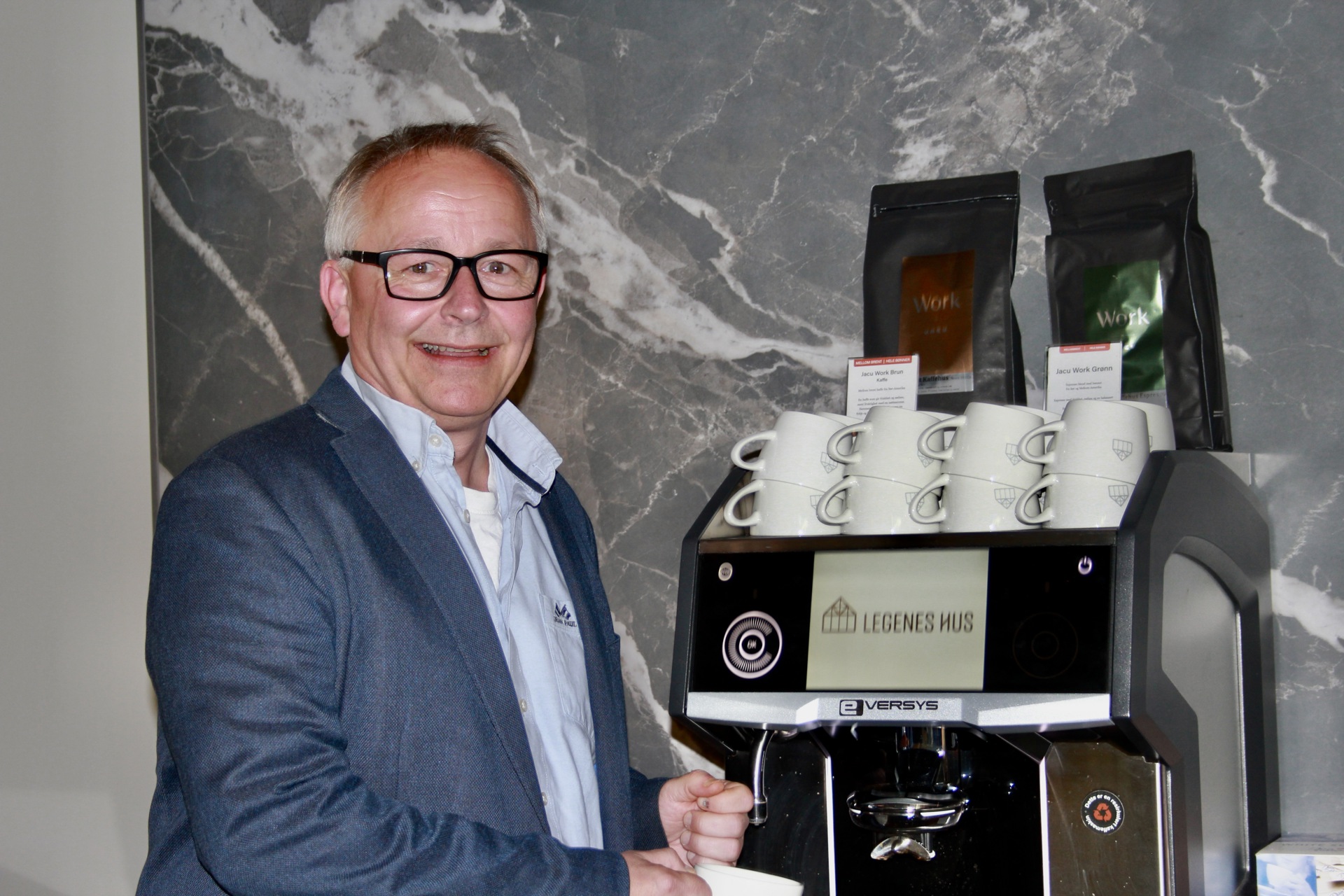 Mann med briller, lyseblå skorte og blå blazer  med kopp i hånden foran kaffemaskin
