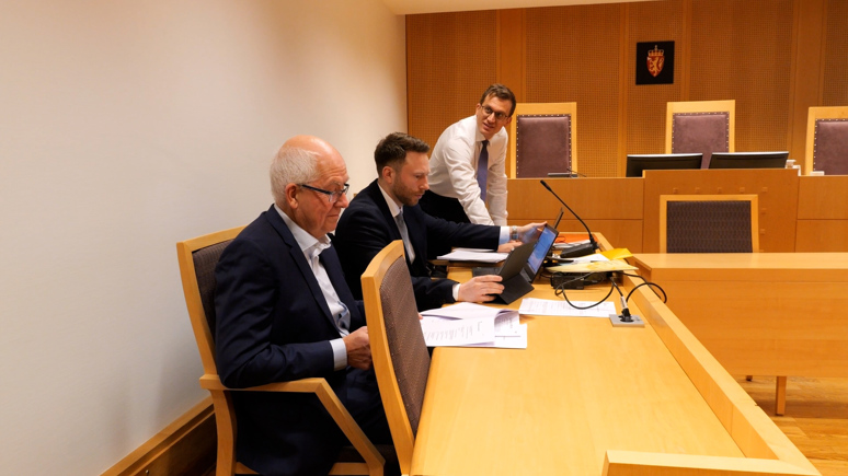Bilde fra en rettssal. Tre menn sitter og står langs en benk til venstre for dommerstolen. Han nærmest kamera sitter, har grått hår og briller, og han har på seg dress. Ved siden av ham sitter en yngre mann med kortklippet, brunt hår og trimmet, kortklippet skjegg. Han har på seg skjorte og dress. Lengst inn i bildet står en mann med lyst, kort hår, briller, hvit skjorte og blått slips. Han lener seg litt frem over benken. 