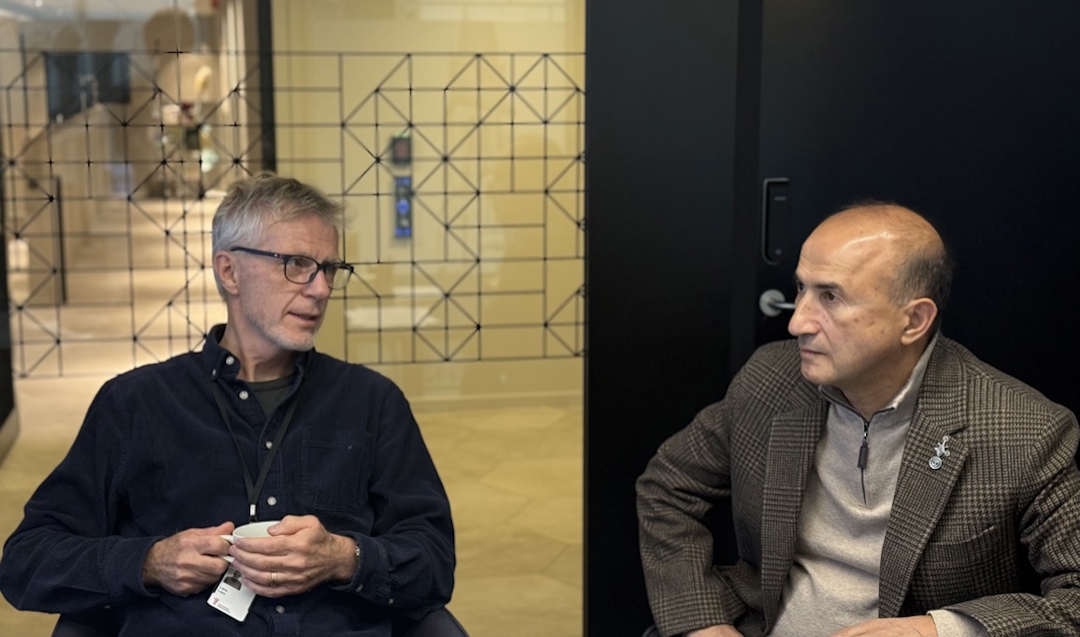 To menn som snakker sammen. Til venstre en mann med grått hår, briller og mørk genser. Til høyre, en mann med brun genser og jakke.