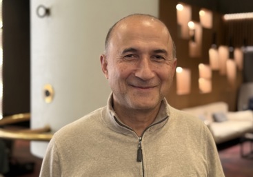 Mann, smilende, beige genser. Lys og hotellobby i bakgrunnen.