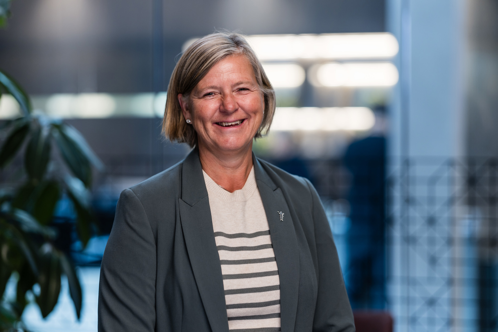 Portrettfoto av president i Legeforeningen, Anne-Karin Rime. Hun står innendørs i et moderne kontormiljø. Hun har kort, lyst hår og er kledd i en grå blazer og en lys genser med horisontale, grå striper. Bakgrunnen er ute av fokus og viser glassvegger, lys fra vinduer og en grønn plante, noe som gir et profesjonelt og rolig miljø.