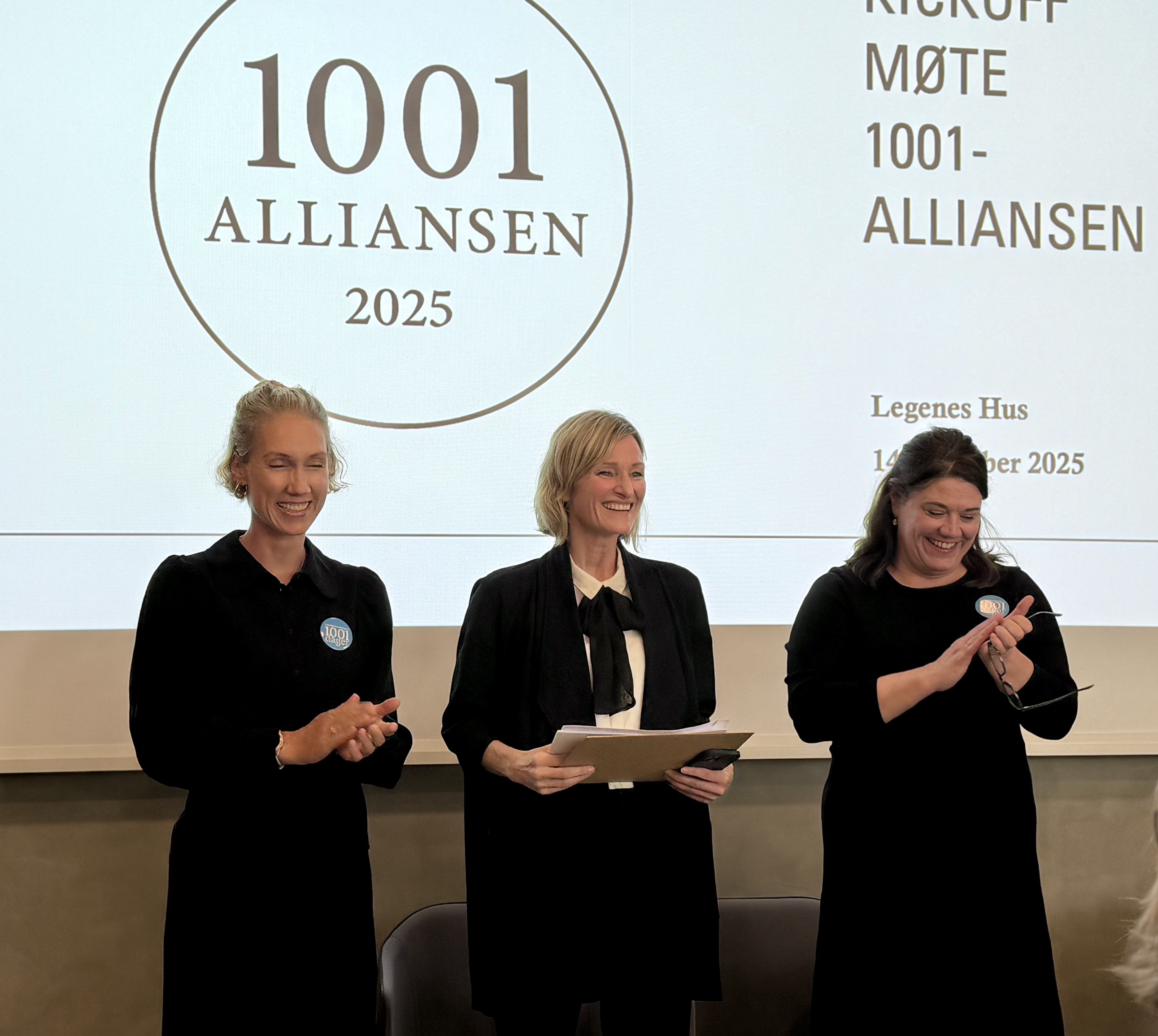 Tre kvinner i m&oslash;rke kl&aelig;r foran en hvit skjerm med teksten 1001-alliansen 2025 