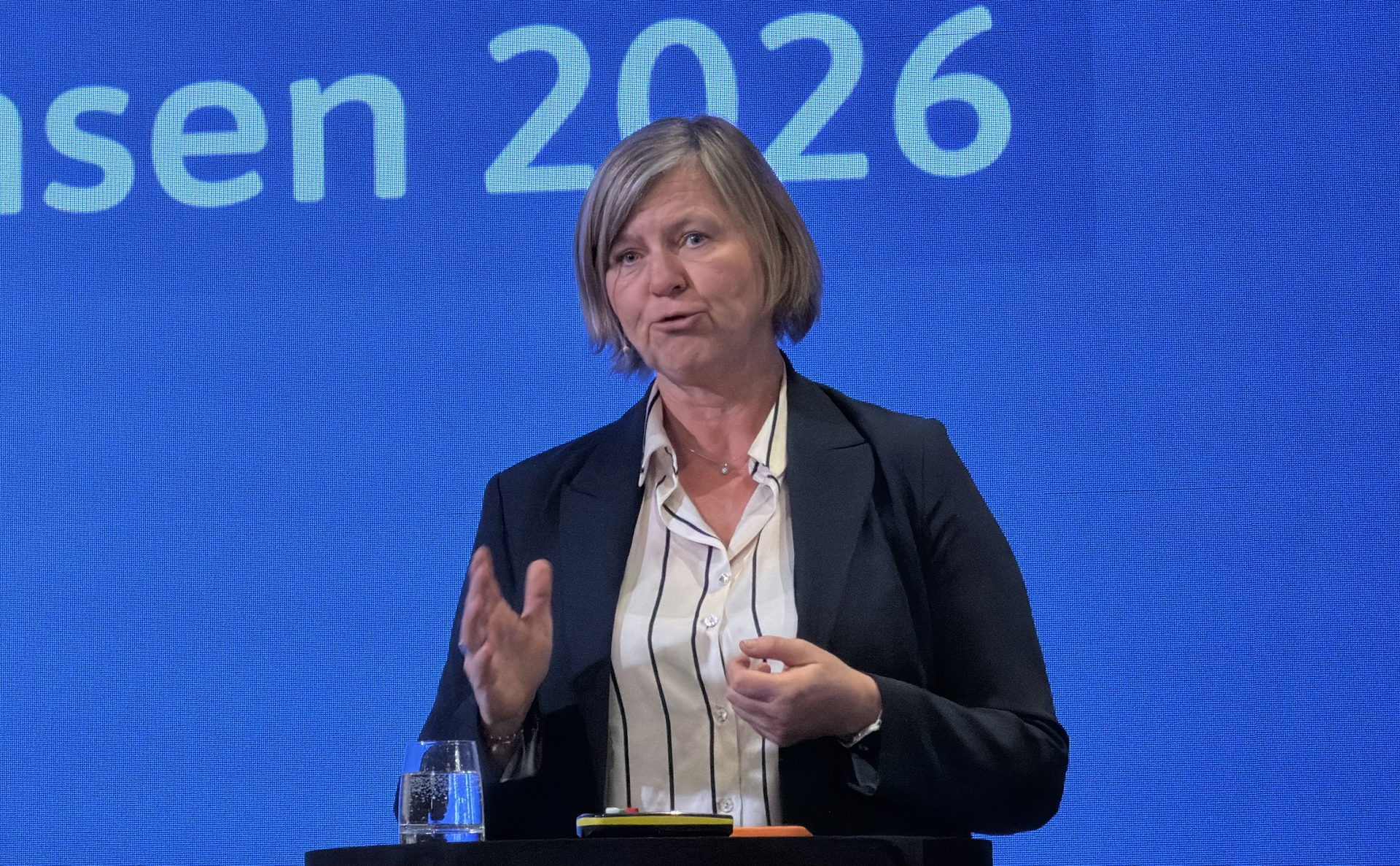 Legepresident Anne-Karin Rime på Spekter sin sykehuskonferanse 2026. Hun står foran en blå bakgrunn med hvit skrift som leser 2026, og vi ser henne fra livet og opp. Hun står foran en talerstol. Hun har lyst hår til haka og med sideskill, blå dressjakke og hvit skjorte med mørkeblå striper. Hun holder på å si noe, og holder ene håndten frem som om hun skjærer i luften med den. 