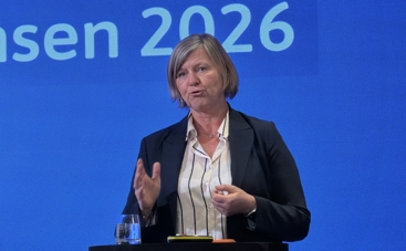 Legepresident Anne-Karin Rime på Spekter sin sykehuskonferanse 2026. Hun står foran en blå bakgrunn med hvit skrift som leser 2026, og vi ser henne fra livet og opp. Hun står foran en talerstol. Hun har lyst hår til haka og med sideskill, blå dressjakke og hvit skjorte med mørkeblå striper. Hun holder på å si noe, og holder ene håndten frem som om hun skjærer i luften med den. 