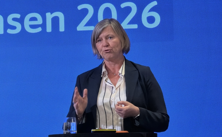 Legepresident Anne-Karin Rime på Spekter sin sykehuskonferanse 2026. Hun står foran en blå bakgrunn med hvit skrift som leser 2026, og vi ser henne fra livet og opp. Hun står foran en talerstol. Hun har lyst hår til haka og med sideskill, blå dressjakke og hvit skjorte med mørkeblå striper. Hun holder på å si noe, og holder ene håndten frem som om hun skjærer i luften med den. 